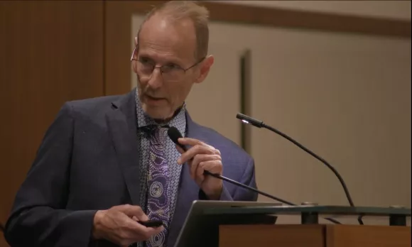 Man in a suit and glasses holding a remote and speaking from a podium.