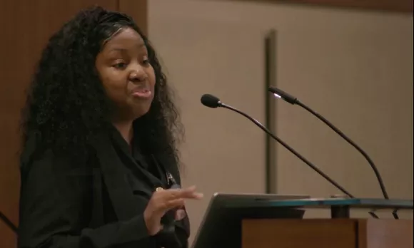 Woman standing at a podium holding a remote and smiling while she speaks.
